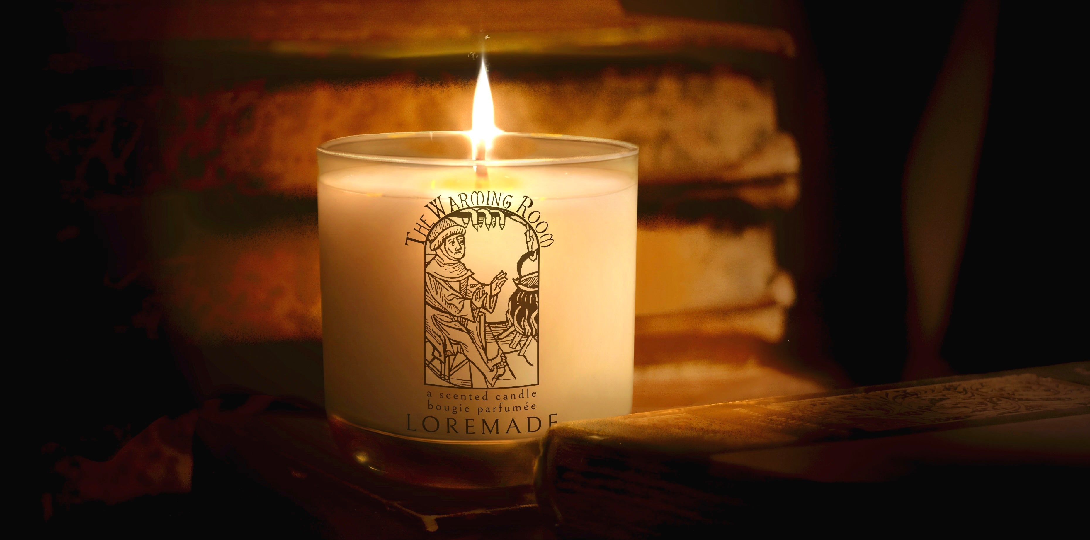 Loremade scented Candle with a label reading The Warming Room on a wooden surface, surrounded by aged manuscripts in a room lit only by the flame of the candle.